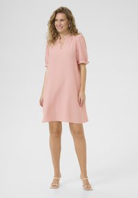 Robe courte rose en tissu texturé, avec un décolleté en V et des manches bouffantes, se terminant au-dessus des genoux. Portée avec des sandales blanches à lanières.