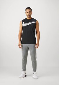 Svart ermeløs T-skjorte med sølvfarget Nike-logo, sammen med lysegrå joggebukser og hvite sneakers. Glatt stoff, atletisk passform.