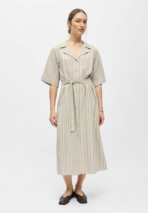 Femme portant une robe rayée beige longue jusqu'aux chevilles avec des manches aux coudes et une ceinture en tissu, associée à des chaussures plates marron foncé.