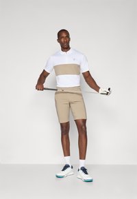 Herre golfantrekk bestående av en hvit polo med en beige stripe, beige shorts og hvite golfsko med marineblå detaljer. Glatt stoff, moderne passform.