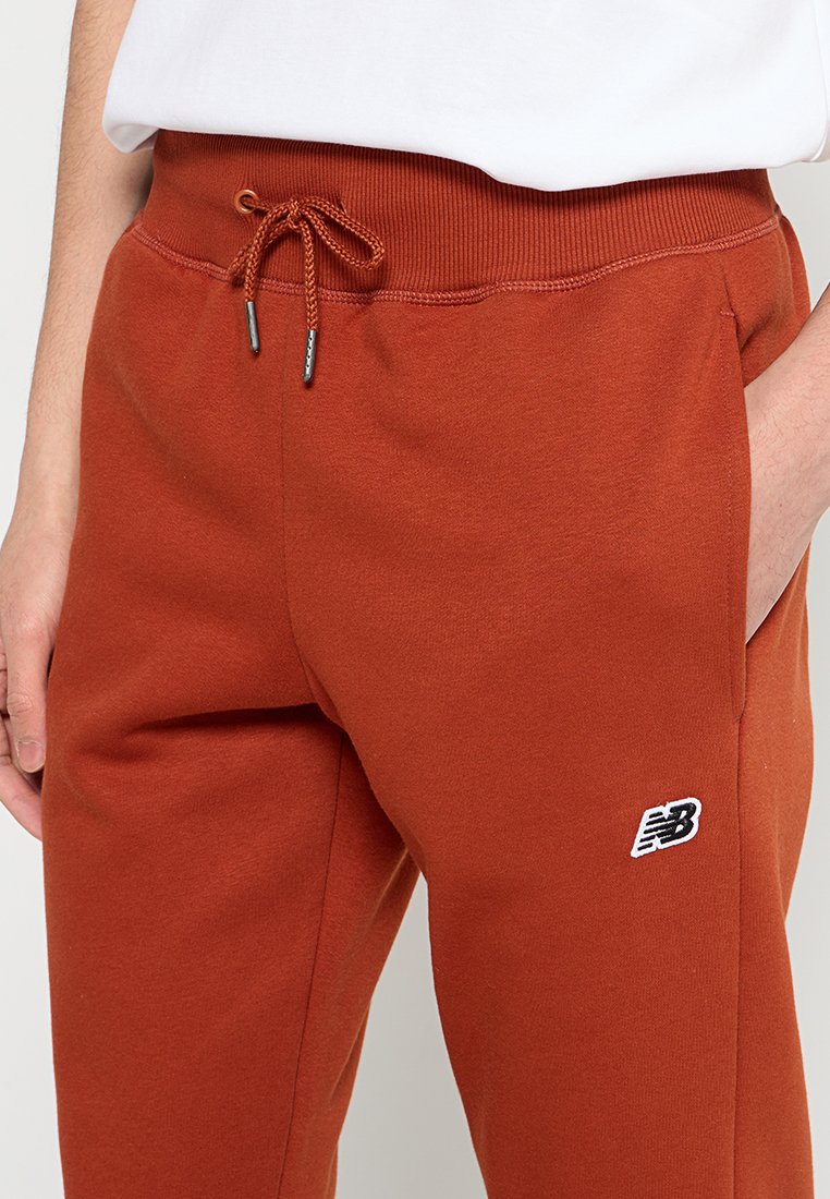 Roestkleurige sweatpants met een aantrekkoord in de taille en het New Balance-logo op de linker bovenbeen, gedragen met een witte shirt ingestopt.