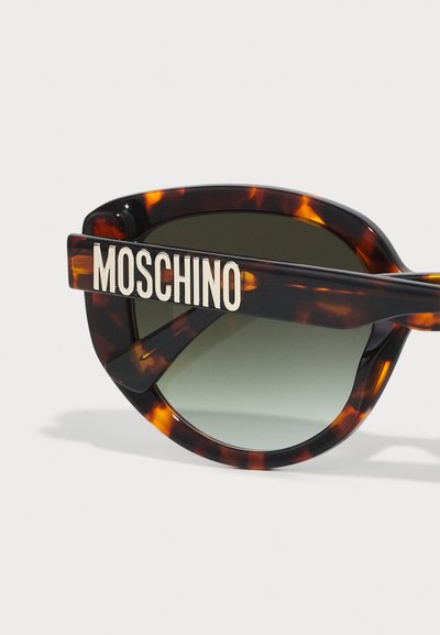Skilpadde-sunglasses med grønne linser. Tykk ramme med en dristig hvit "Moschino"-logo på armen. Glatt tekstur og oversized form.