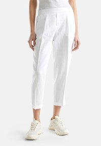 Białe lniane spodnie o luźnym kroju, z plisowanym wzorem, elastycznym paskiem i podwiniętymi mankietami, w zestawieniu z jasnymi sneakersami.