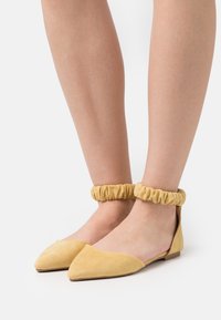 Mocasines de ante amarillo con punta afilada y tiras elásticas en el tobillo. Los zapatos tienen un tacón bajo y una textura suave.