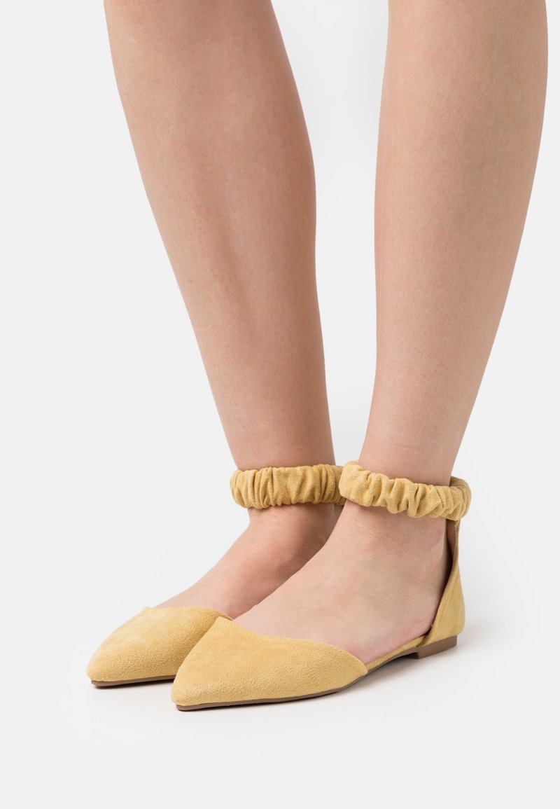 Mocasines de ante amarillo con punta afilada y tiras elásticas en el tobillo. Los zapatos tienen un tacón bajo y una textura suave.