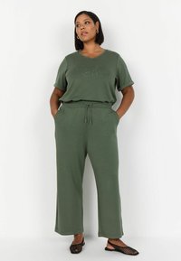 Completo coordinato color verde oliva con scollatura a V, maniche corte e pantaloni in stile jogger. I pantaloni hanno una vita con cordoncino elasticizzato e tasche laterali.