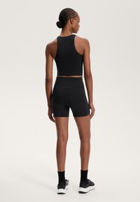 Femme avec des cheveux tressés portant un crop top sans manches noir, un short de vélo taille haute, des chaussettes noires et des baskets, se tenant devant un fond neutre.