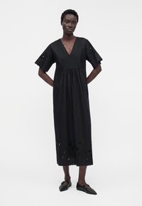 Femme aux cheveux courts portant une robe midi noire à col en V avec un ourlet festonné et des motifs floraux découpés, associée à des ballerines noires pointues.