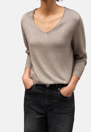 Kobieta ubrana w beżowy sweter z dekoltem w serek i czarne, damskie, wysokie jeansy z rękami w kieszeniach na jednolitym tle.