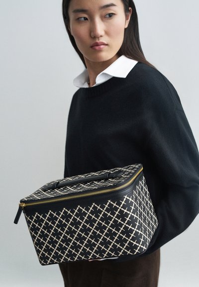 Svart och krämfärgad mönstrad väska med rektangulär form, strukturerad yta och guldfärgad dragkedja, hålls av en person i en svart tröja.