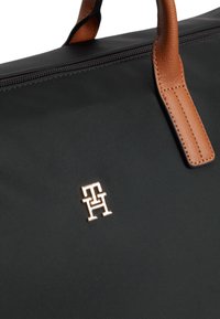 Μαύρη υφασμάτινη τσάντα με καφέ δερμάτινα χερούλια και μικρό μεταλλικό λογότυπο που συνδυάζει τα γράμματα T και H κοντά σε μαύρο φερμουάρ.