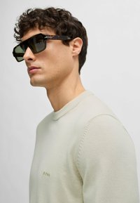 Sonnenbrille mit rechteckigem Rahmen im braunen Schildpatt-Muster, grünen Gläsern und dünnen Metallakzenten. Modell trägt einen hellbeigen Pullover.