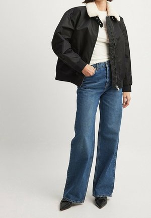 Personne portant une veste bomber noire avec col en polaire blanc, chemise blanche, jean bleu à jambes larges et chaussures noires à bout pointu, debout avec une main dans la poche.