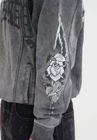 Manica di giacca di jeans grigia con design in stile tatuaggio bianco e nero che presenta una rosa, un'ala, un cuore e fiamme.