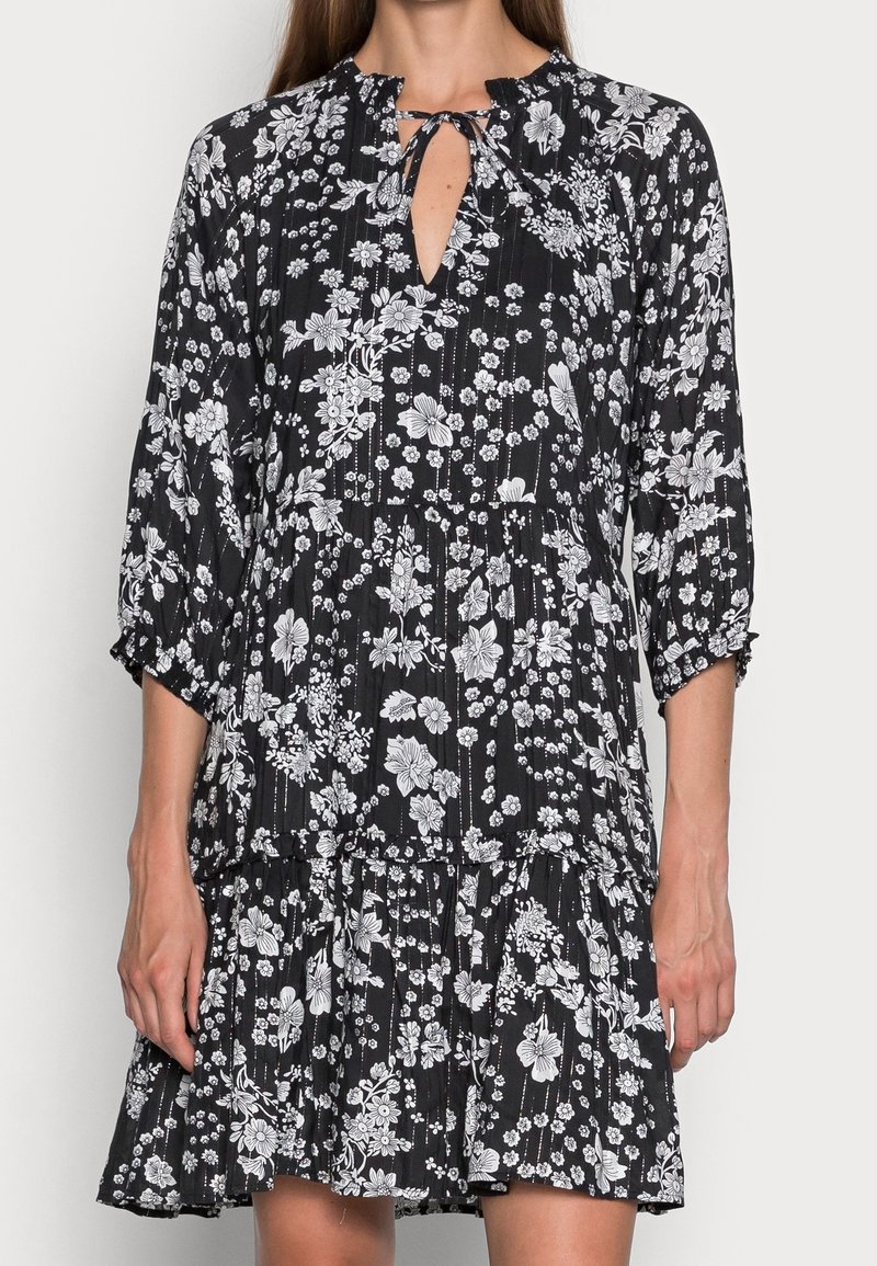 Robe noire à motif floral blanc, manches trois-quarts, encolure avec ouverture en forme de goutte nouée par un nœud, et jupe à volants.