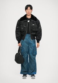 Chaqueta bomber negra con cuello de piel sintética y múltiples bolsillos, combinada con pantalones cargo sueltos y brillantes en color teal y un bolso negro. Zapatillas blancas completan el atuendo.