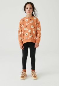 Orange sweatshirt med vit och svart blommönster, svarta leggings och bruna sneakers med rosa detaljer på ett barn som står på en neutral bakgrund.