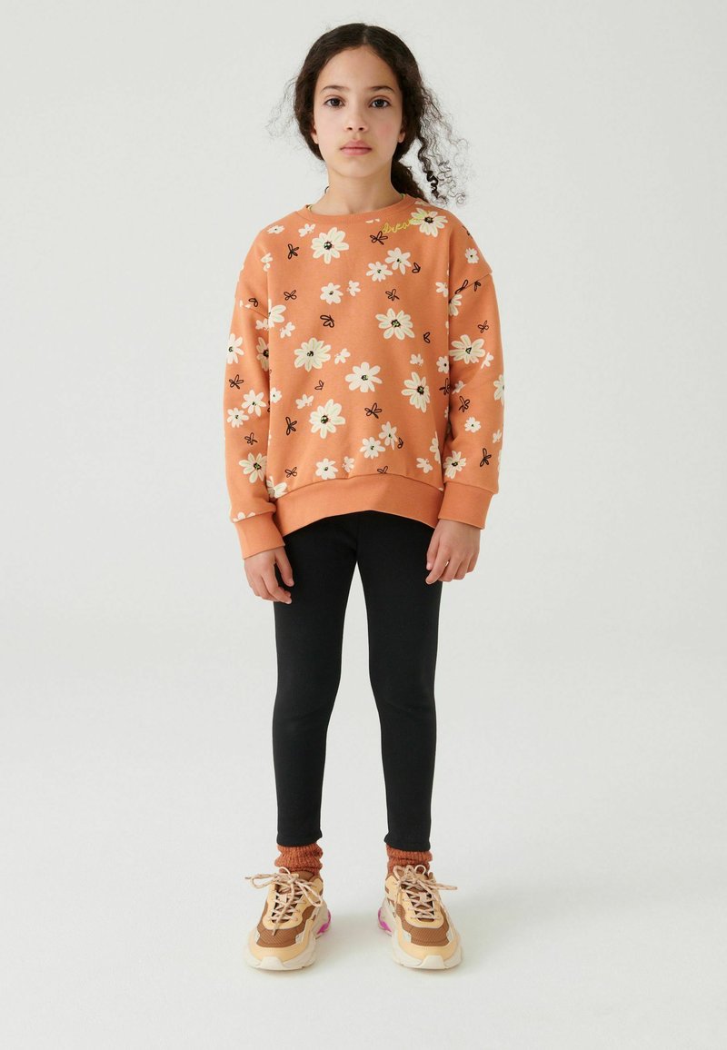 Orange sweatshirt med vit och svart blommönster, svarta leggings och bruna sneakers med rosa detaljer på ett barn som står på en neutral bakgrund.