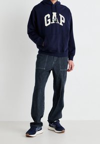 Felpa blu navy con logo "GAP", abbinata a pantaloni a righe blu navy. Include una tasca a marsupio e polsini a coste. Indossata con sneakers blu navy.