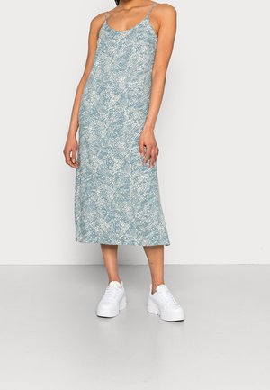 Nainen, joka pukeutuu sinivalkoiseen kukalliseen keskipitkään liivimekkoon, jossa on ohuet olkaimet, ja valkoisiin korollisiin tennareihin, seisoo yksiväristä taustaa vasten.