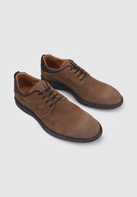 Chaussures en daim marron avec un embout arrondi, des lacets noirs fins et une partie supérieure sombre contrastante. La semelle est noire avec un léger relief.