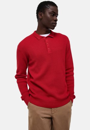 Homme regardant sur le côté, portant un polo rouge texturé à manches longues et un pantalon beige, les mains dans les poches, sur fond blanc.