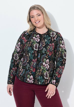 Smilende kvinde med blondt hår iført en sort jakke med blomsterprint og mørkerøde bukser, stående mod en hvid baggrund.