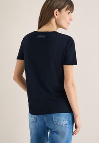 Femme portant un T-shirt noir avec le texte « CURIOUS » sur le haut du dos et un jean en denim bleu, debout contre un mur clair uni.