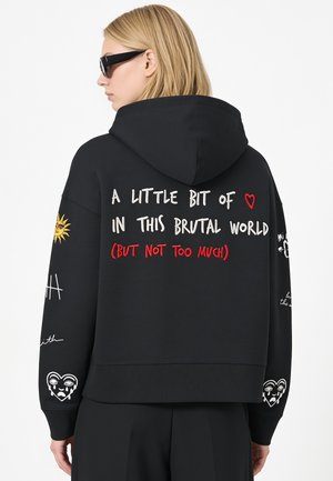 Blonde Person trägt schwarzen Hoodie mit weißem und rotem Text, Sonnen- und Herzgrafiken, schaut zur Seite vor schlichtem weißem Hintergrund.