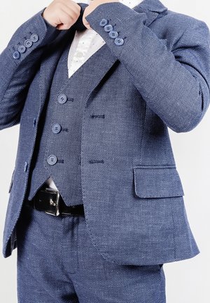 Dziecko poprawiające granatową teksturowaną marynarkę i kamizelkę na białej koszuli, ukazując pasek i detale guzików.