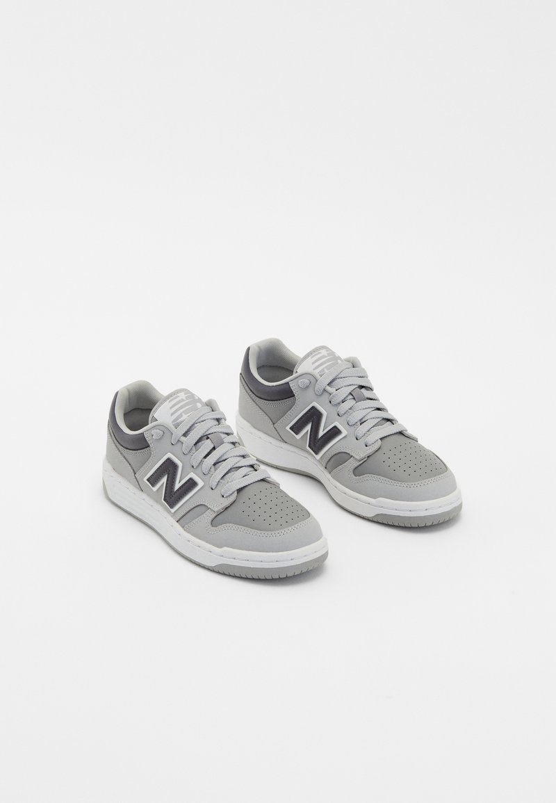 Paire de baskets New Balance grises avec semelles et lacets blancs, zone des orteils perforée, et logo noir "N" sur les côtés.