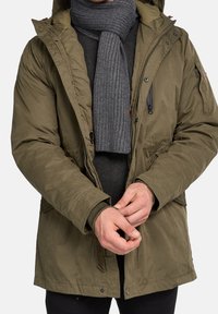 Olivgrüner isolierter Parka mit Kapuze, Reißverschluss und Druckknopfverschlüssen sowie einem gerippten grauen Schal. Strukturiertes Material mit Seitentaschen.