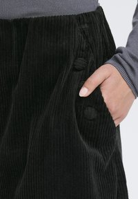 Pantaloni negri din catifea cu un suprafață texturată, două nasturi decorativi din țesătură și buzunare laterale. Vedere de aproape a zonei buzunarului.