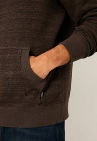 Dunkelbrauner, strukturierter Hoodie mit gerippten Bündchen an Ärmeln und Bund, versehen mit einer seitlichen Fronttasche. Etikett an der Naht sichtbar.