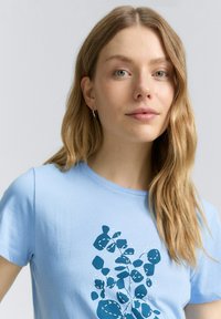 Camiseta de algodón azul claro con un gráfico inspirado en plantas en azul oscuro y detalles en blanco. Manga corta y escote redondo.