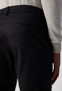 Black trousers featuring a flat back pocket with a button closure, made of smooth fabric, paired with a light gray sweater.