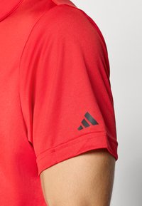 Červené športové tričko s krátkym rukávom nosené na ruke, s čiernym logom Adidas vytlačeným blízko okraja rukávu.