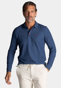 Donkerblauwe polo met lange mouwen en een kraag, voorzien van een roze knoop en een klein rood logo op de borst. G gedragen met beige broek.