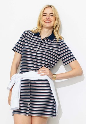 Femme blonde souriante, portant une robe boutonnée à manches courtes rayée bleu marine, blanc et rouge avec un pull blanc noué autour de sa taille.