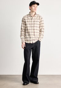 Camicia checked in beige e marrone, abbinata a jeans scuri. La camicia ha colletto e maniche lunghe; cappellino nero indossato.