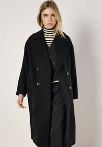 Schwarzer Wollmantel mit einem doppelt knöpfbaren Design, großen Revers und zwei Knöpfen. Getragen über einem schwarz-weißen gestreiften Rollkragenpullover.