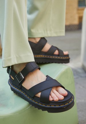 Sorte sandaler med brede krydsremme, justerbar spænde, tyk tekstureret sål og gule syninger, hvilende på en grøn overflade.