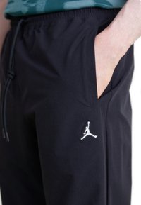 Mann trägt schwarze Jogginghosen mit einer Hand in der Tasche, versehen mit einem weißen Jumpman-Logo auf dem Oberschenkel und einem elastischen Bund mit Kordelzug.