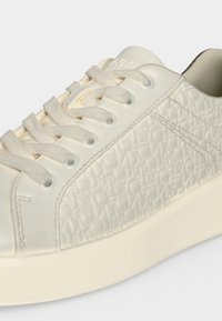 Gros plan d'une basket blanc cassé avec un panneau latéral texturé présentant des motifs répétitifs en relief et des lacets plats en tissu.