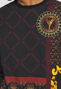 Langærmet sort skjorte med røde og gyldne mønstre, herunder en logo, geometriske detaljer og blomsterdesign langs kanten og ærmerne.