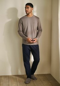 Maglia marrone a maniche lunghe con tasca, abbinata a pantaloni blu scuro. La modella è in piedi su un pavimento in legno, indossa pantofole grigie con un motivo a strisce.