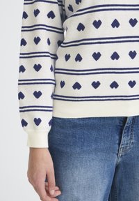 Pull en tricot crème avec des rayures horizontales bleu marine et des motifs en forme de cœur ; poignets et ourlet côtelés ; associé à un jean en denim.