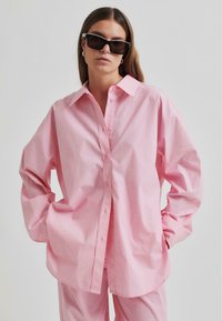 Camicia in cotone rosa oversize con colletto a punta, bottoni frontali e maniche lunghe, abbinata a pantaloni coordinati. Occhiali da sole neri completano il look.
