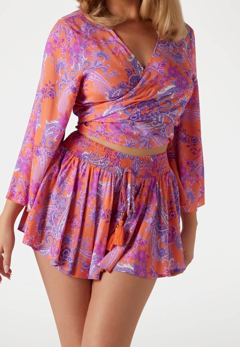 Femme portant un top cache-cœur floral orange et violet et une mini-jupe fluide assortie avec un détail de nouage à pompons, coupée au niveau du ventre.