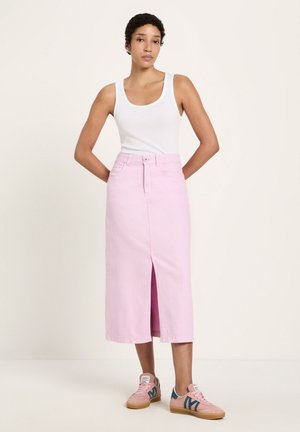 Woman standing with hands behind back wearing white tank top, pink midi skirt with front slit, and pink sneakers with blue accents.